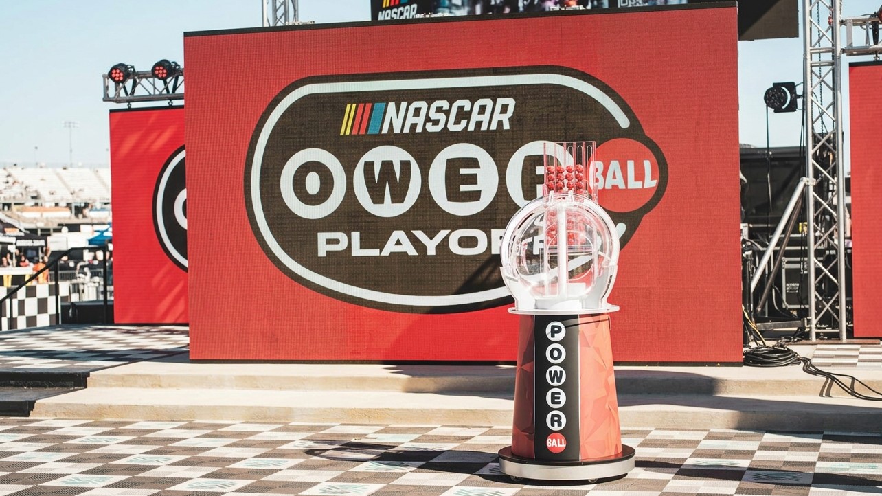 Powerball Playoff drawing machine filled with red lottery balls on a stage in front of a NASCAR-branded screen